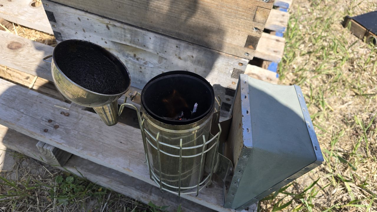 A copper bee smoker with a wire heat-guard cage, lit with a small flame visible inside, sitting on a weathered wood hive box next to a brass funnel-scoop full of dark charred fuel and a galvanized metal box.